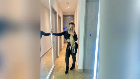 Woman with long blonde braids in a black crop top and leggings taking a mirror selfie in a modern hallway.