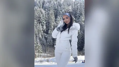 Smiling woman in white winter ski outfit and headband posing in a snowy pine forest landscape.