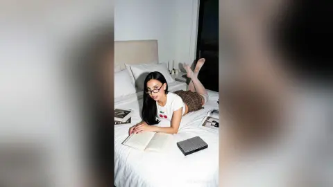 Young woman in white t-shirt reading a book on a white bed in a bright, cozy bedroom setting.