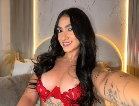 Smiling woman with long dark hair wearing a red lace bra in a cozy, warmly lit bedroom setting.