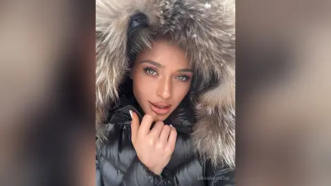 Beautiful woman in a black puffer jacket and large faux fur hood smiling in a snowy winter setting.