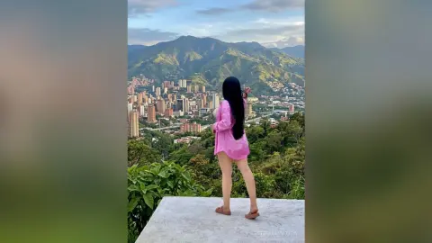 Woman in pink shirt overlooking a scenic mountain city view from a lush green hilltop at sunset.