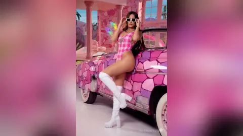 Glamorous woman in a pink patterned bodysuit and white boots posing on a pink vintage car in a dreamy pastel setting.