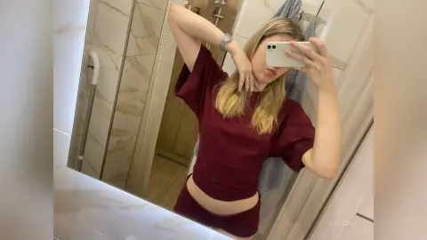 Young woman in a maroon t-shirt taking a casual mirror selfie in a modern marble bathroom.