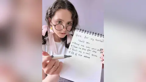 Young woman in glasses holding a notebook with funny teacher quiz notes in a soft, bright indoor setting.