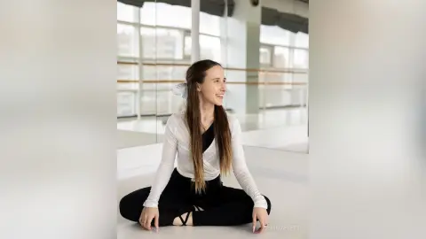 Smiling woman in white long-sleeve shirt and black leggings sitting in a bright, modern dance studio.
