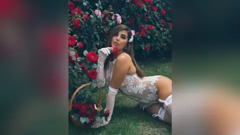 AllishaCoral — Woman in white lace bodysuit sitting in a rose garden holding a basket of red flowers in a romantic mood.