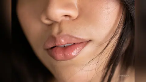 Close-up of a woman with glossy pink lips and natural makeup in soft, warm lighting.