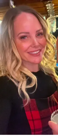Smiling blonde woman in a black and red plaid outfit enjoying a cozy evening at a dimly lit restaurant.