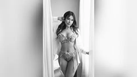 Elegant woman in floral lace lingerie and a white robe posing in a bright, minimalist indoor setting.