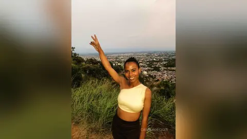 Smiling young woman in a yellow crop top making a peace sign on a grassy hill overlooking a city landscape.