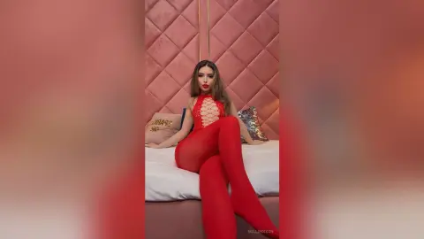 Glamorous woman in a red lace bodysuit sitting on a bed against a pink quilted headboard.