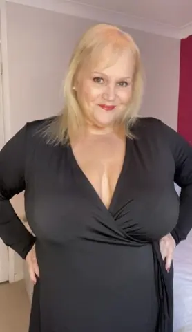 Busty_Madeline poses in a black long-sleeved wrap dress, showing cleavage and her blonde hair against a plain wall background.