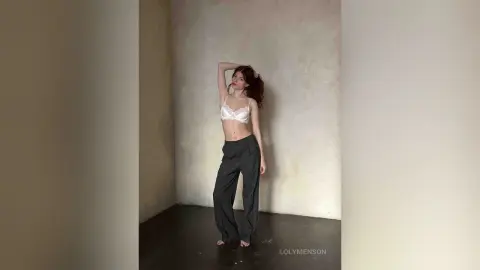 Young woman in a white lace bralette and grey wide-leg trousers posing in a minimalist studio setting.