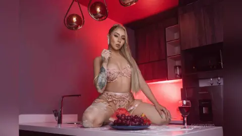 Blonde woman in lace lingerie posing in a moody, red-lit modern kitchen setting.
