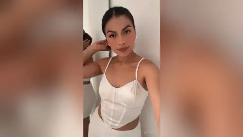 Young woman in a white corset top and matching skirt posing in a bright, minimalist indoor setting.