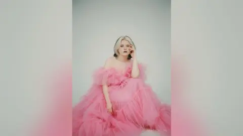 Blonde woman in a voluminous pink tulle gown posing elegantly against a minimalist light background.