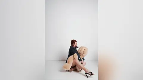 AryaSin sits on a white floor, wearing a black top and hugging a large tan teddy bear against her body.