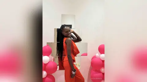 Smiling Black woman in a vibrant red jumpsuit posing in a bright room with pink balloons.
