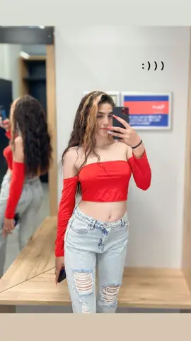 Young woman in a red off-the-shoulder crop top and ripped light wash jeans taking a mirror selfie.