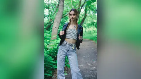 Young woman in a black leather jacket and striped crop top posing on a lush green forest path.