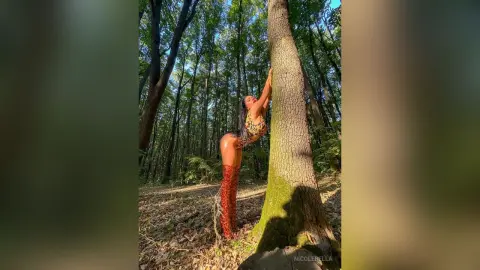 NicoleBella arches her back against a large tree trunk in a forest, wearing a leopard print bodysuit and red thigh-high boots.