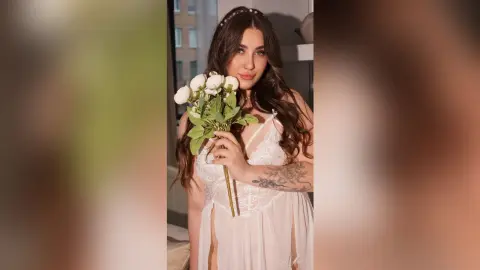KeiraHarts holds a bouquet of white roses while wearing a white lace teddy that opens in the front to show her stomach and lower body.