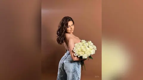 Elegant woman in denim jeans holding a large bouquet of white roses against a warm brown studio background.
