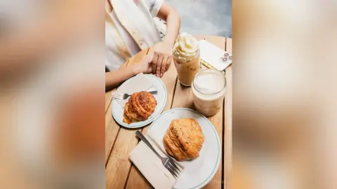 TefyRusso sits at a wooden table with two croissants and two iced coffee drinks, wearing a white sleeveless top.