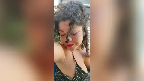 Smiling woman with curly hair wearing a black polka dot sundress in a bright outdoor setting.
