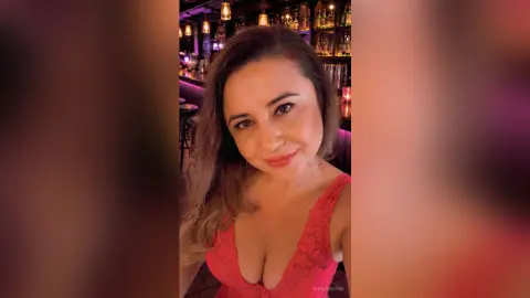MaliaQuinn poses in a pink lace top with a deep cleavage, looking at the camera in a dimly lit bar setting.