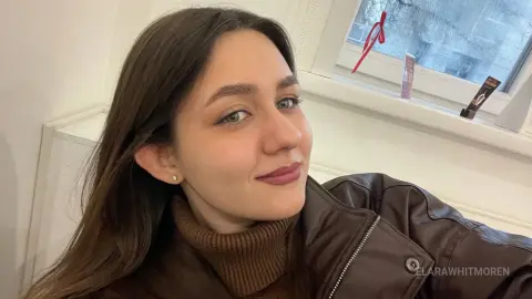 Smiling young woman in a brown leather jacket and turtleneck sweater posing indoors near a window.