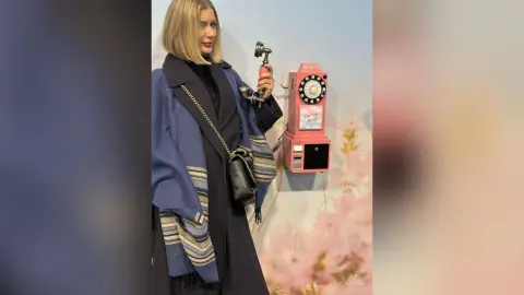 Stylish blonde woman in a blue and black coat holding a vintage pink rotary telephone against a floral backdrop.