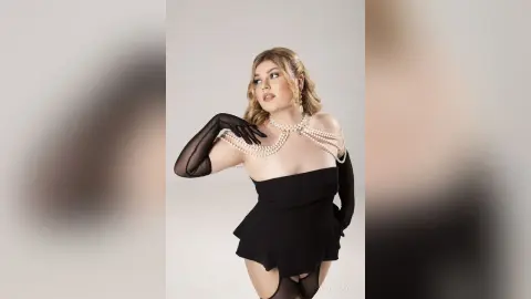 Elegant woman in a black mini dress and long black gloves wearing a pearl necklace against a white studio background.