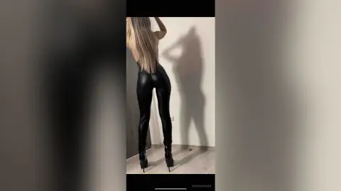 Blonde woman in black leather leggings and high heels posing against a minimalist wall.
