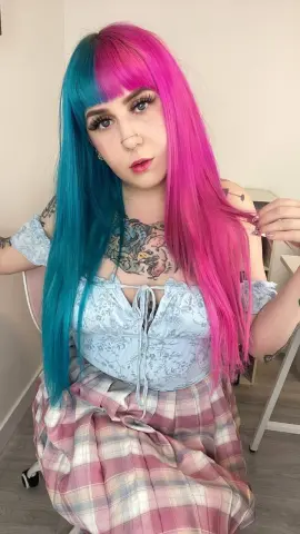 Young woman with split dye pink and blue hair wearing a light blue floral top and plaid skirt in a bright room.