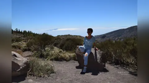 Woman in light blue casual outfit sitting on a rock in a sunny mountain landscape under a clear blue sky.