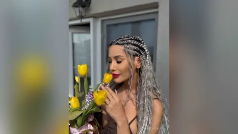 Woman with long silver braids holding yellow tulips in a bright outdoor setting with a joyful mood.
