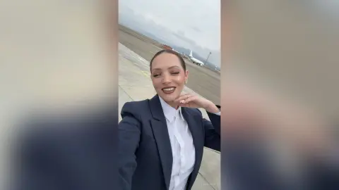Smiling woman in a professional navy blazer and white shirt standing on an airport tarmac under a cloudy sky.