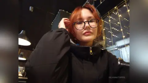 Redheaded woman in a black puffer jacket and glasses posing in a dimly lit room with fairy lights.