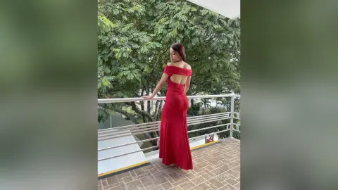 KimRece stands on a balcony wearing a long, tight red dress that shows her bare back and shoulders.