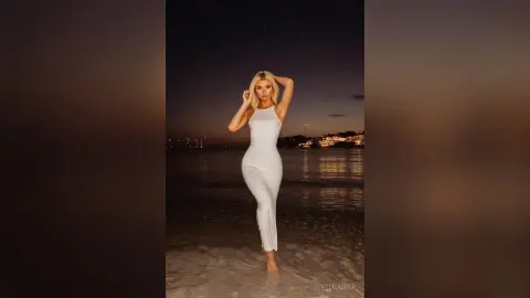 Blonde woman in a sleek white maxi dress posing on a beach at night under a dark sky.