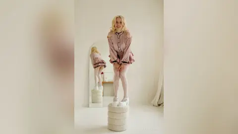LynGoodhart poses in a pink ruffled dress and white platform shoes while standing on a white pedestal in a bright room.