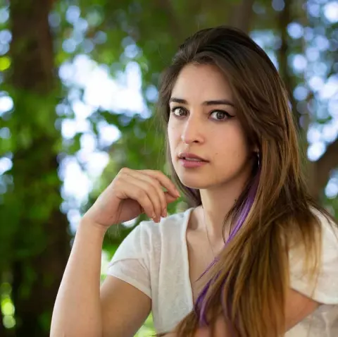 Young woman with long brown hair wearing a white v-neck shirt in a lush green forest setting.