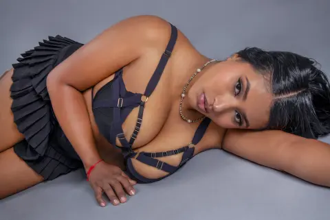 Beautiful woman in black lingerie and pleated skirt posing on a grey background with a moody aesthetic.