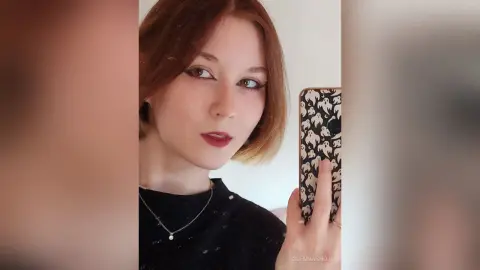 Redheaded woman in a black top taking a selfie with a spooky ghost pattern phone case in a bright room.