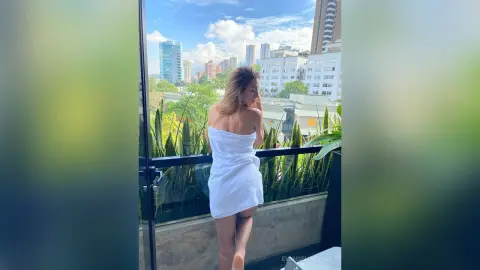 Woman in a white towel standing on a high-rise balcony overlooking a sunny city skyline.