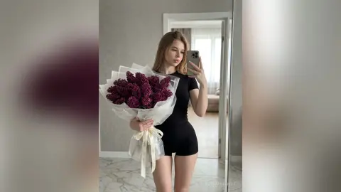 Young woman in a black bodysuit holding a large bouquet of purple lilacs in a bright modern room.