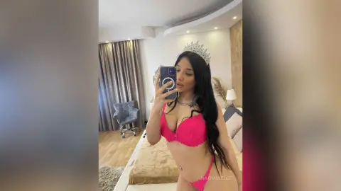 Young woman in pink lace lingerie and a silver tiara taking a mirror selfie in a bright bedroom.
