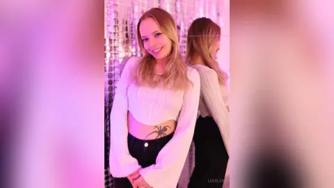 LucyLoy smiles in a white long-sleeve crop top and black pants, showing off a large spider tattoo on her stomach.